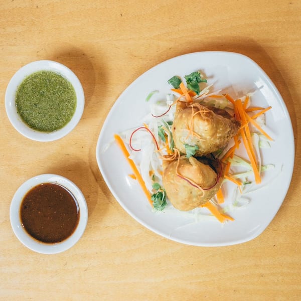 Two golden-brown samosas on a white plate with shredded cabbage, carrots, and two dipping sauces.