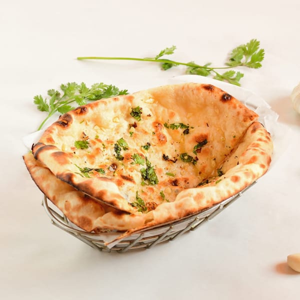 Freshly baked garlic naan bread, garnished with cilantro, in a metal basket on a white surface.